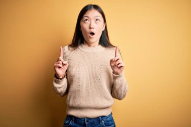 Sarı arka plan üzerine günlük süveter giyen genç, güzel Asyalı kadın şaşırmış ve şaşırmış bir şekilde yukarı bakıyor ve parmaklarıyla işaret ediyor ve kollarını kaldırıyordu..