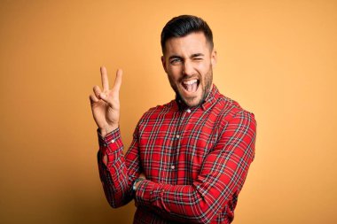 Izole sarı arka plan üzerinde duran rahat gömlek giyen genç yakışıklı adam zafer işareti yapıyor kameraya göz kırparak mutlu yüz ile gülümseyerek. İki numara.
