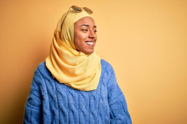 Sarı başörtüsü ve güneş gözlüğü takan Afro-Amerikalı genç bir bayan yüzünde gülümseme, doğal bir ifade ile diğer tarafa bakıyor. Kendinden emin gülen.