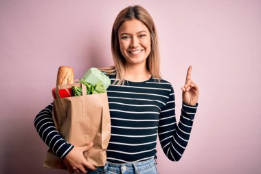 Young beautiful woman holding paper bag with purchase over isolated pink background surprised with an idea or question pointing finger with happy face, number one