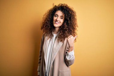 Kıvırcık saçlı, piercingli, sıradan tişörtlü, mutlu yüzüyle gülümseyen ve baş parmağıyla yüzünü gösteren genç, güzel esmer kadın..