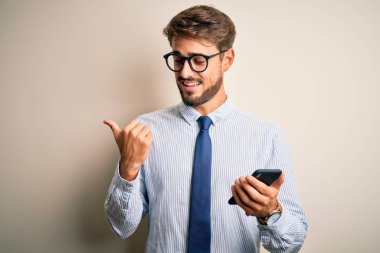 Genç işadamı beyaz arka plan üzerinde akıllı telefon kullanarak konuşuyor ve gülümseyen bir gülümsemeyle baş parmağıyla onu işaret ediyor.