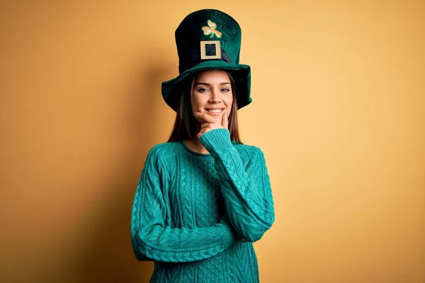 Young beautiful brunette woman wearing green hat with clover celebrating saint patricks day looking confident at the camera smiling with crossed arms and hand raised on chin. Thinking positive.