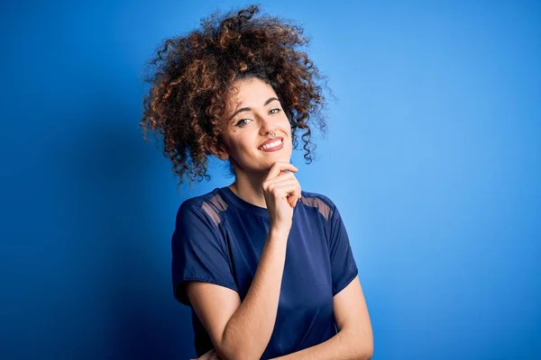 Young beautiful woman with curly hair and piercing wearing casual blue t-shirt looking confident at the camera with smile with crossed arms and hand raised on chin. Thinking positive.