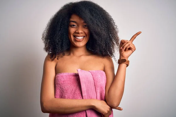 Young african american woman with afro hair wearing a body towel after beauty care shower with a big smile on face, pointing with hand and finger to the side looking at the camera.