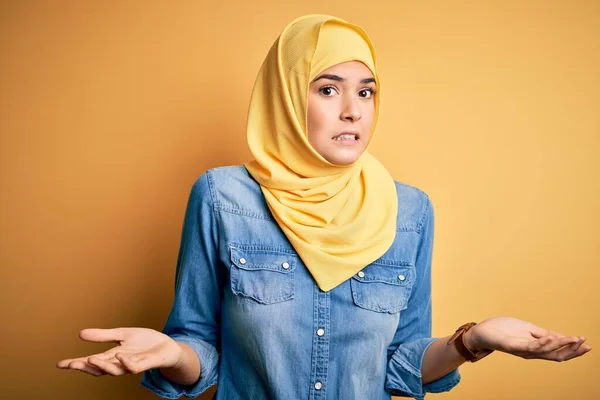 孤立した黄色の背景の上に立つイスラム教徒のヒジャーブを身に着けている若い美しい少女は 腕や手を上げて混乱表現 疑いの概念 — ストック写真