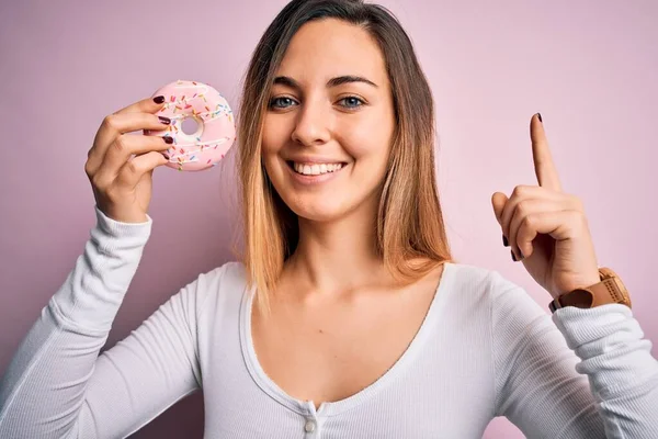 若いです美しいですブロンド女性とともに青目保持ピンクドーナツ上の隔離された背景驚きでAアイデアや質問指差し指とともに幸せな顔 ナンバーワン — ストック写真