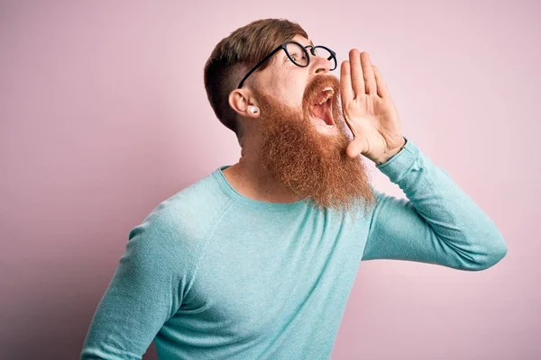 Handsome Irish redhead man with beard wearing glasses over pink isolated background shouting and screaming loud to side with hand on mouth. Communication concept.