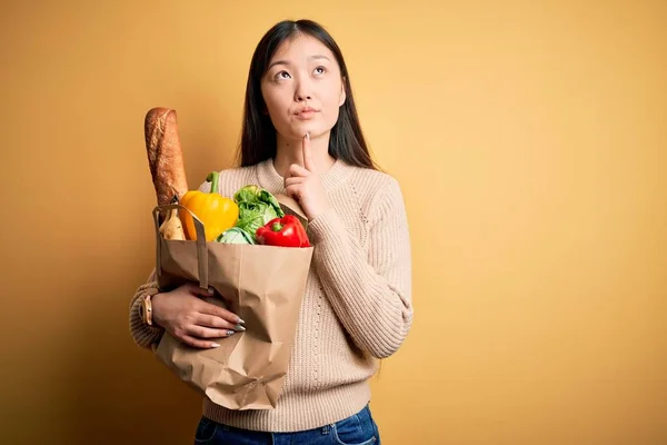 年轻的亚洲女人拿着一袋袋新的健康食品放在黄色孤立的背景下 满腹狐疑 仰起头纳闷 — 图库照片