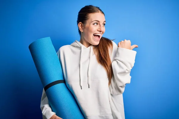 年轻美丽的红头发女运动员拿着瑜伽垫在蓝色的背景上 手指着身体侧面 笑容满面 — 图库照片