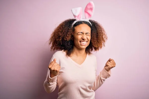 ピンクの背景の上にウサギの耳を身に着けているアフロの髪を持つ若いアフリカ系アメリカ人の女性は非常に満足し 腕を上げて勝者のジェスチャーを行う興奮しました 笑顔と成功のために叫んで お祝いのコンセプト — ストック写真