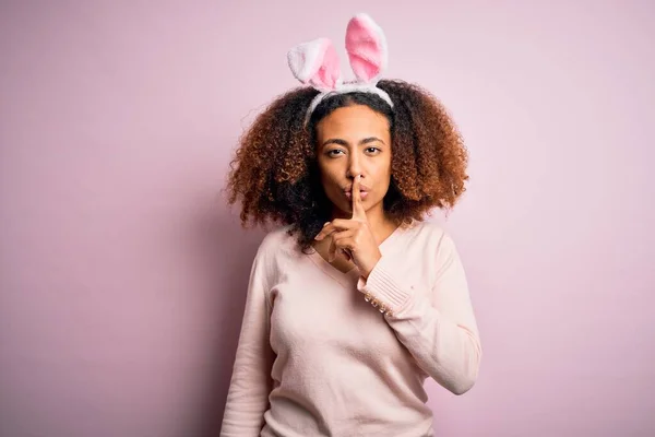 年轻的非洲裔美国女人 一头黑发 粉红的背景上戴着小兔子耳朵 要求保持安静 手指放在嘴唇上 沉默和秘密概念 — 图库照片