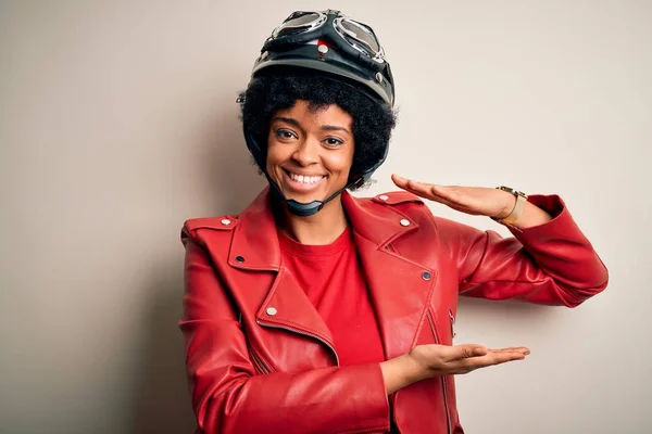 Young African American afro motorcyclist woman with curly hair wearing motorcycle helmet gesturing with hands showing big and large size sign, measure symbol. Smiling looking at the camera. Measuring concept.