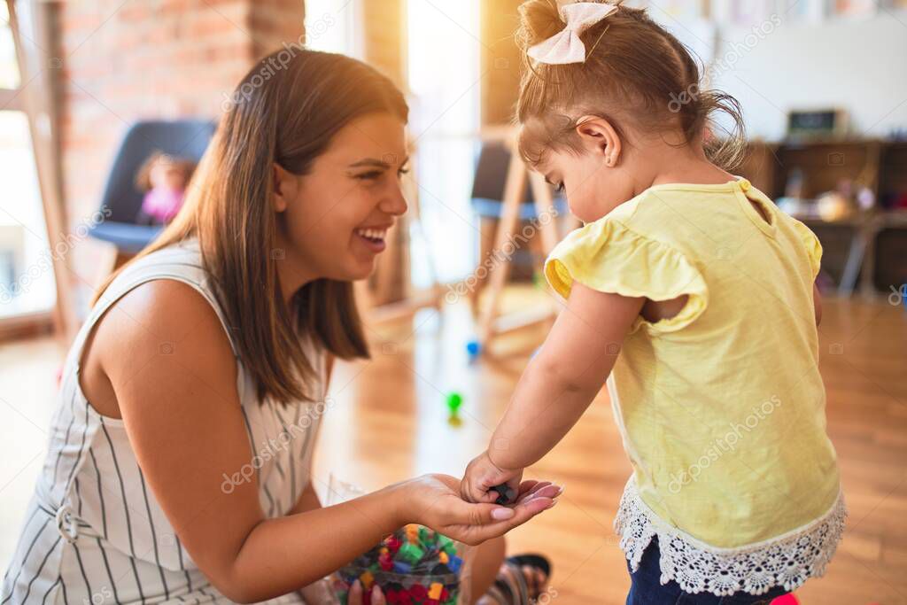 Young beautiful teacher and toddler playing with small building blocks toy at kindergarten