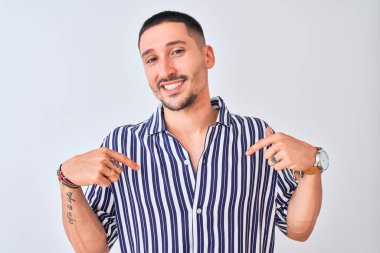 Deniz çizgili gömlek giyen genç yakışıklı adam izole edilmiş arka planda duruyor. Yüzünde bir gülümsemeyle, gururlu ve mutlu bir şekilde kendini işaret ediyor..