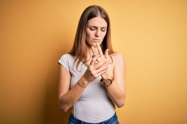 Güzel, kızıl saçlı bir kadın izole edilmiş sarı arka planda günlük tişört giyiyor. Ellerinde ve parmaklarında ağrı var. Eklem iltihabı.