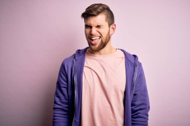 Sakallı, mavi gözlü, pembe arka planda mor eşofman üstü giyen, kameraya seksi bir ifadeyle bakan, neşeli ve mutlu yüzlü genç bir sarışın..