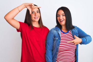 İzole edilmiş beyaz arka planda sıradan kıyafetler giyen genç ve güzel kadınlar gülümseyerek mutlu yüzlü elleri ve parmakları olan bir çerçeve yapıyorlar. Yaratıcılık ve fotoğrafçılık kavramı.