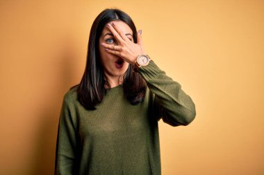 Mavi gözlü genç esmer kadın sarı arka planda yeşil bir süveter giyiyor. Şok olmuş bir şekilde yüzünü ve gözlerini kapıyor. Utanmış bir ifadeyle parmaklarının arasından bakıyor..