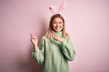 Young beautiful woman wearing easter rabbit ears standing over isolated pink background smiling and looking at the camera pointing with two hands and fingers to the side.