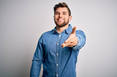 Young handsome blond man with beard and blue eyes wearing casual denim shirt smiling friendly offering handshake as greeting and welcoming. Successful business.