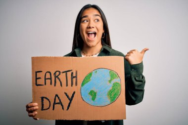 Young beautiful asian woman holding banner asking for earth and enviroment conservation pointing and showing with thumb up to the side with happy face smiling
