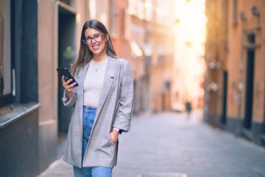 Mutlu ve kendinden emin gülümseyen genç ve güzel bir kadın. Yüzünde gülümsemeyle şehirde akıllı telefon kullanarak durmak.