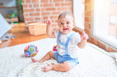 Güzel bebek battaniyenin üzerinde oturuyor. Anaokulunda elleri havada gülümsüyor.