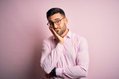 Pembe arka planda duran zarif gömlek ve gözlük takan yakışıklı genç adam kollarını kavuşturarak depresyondan sıkılmış ve yorgun görünüyor..