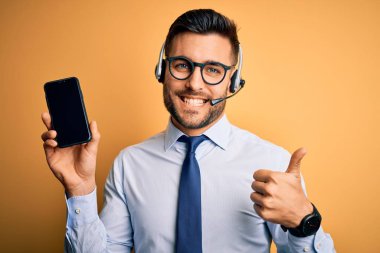 Çağrı merkezinden gelen müşteri hizmetleri kulaklığı olan bir işletmeci akıllı telefon ekranını büyük bir gülümsemeyle gösteriyor. Tamam işareti yap, parmaklarınla başparmağını kaldır, mükemmel işaret.