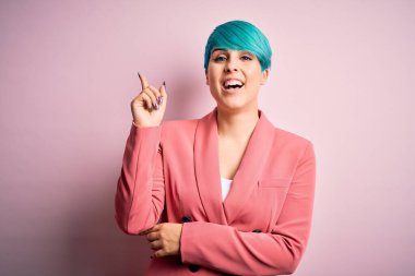 Young beautiful businesswoman with blue fashion hair wearing jacket over pink background with a big smile on face, pointing with hand and finger to the side looking at the camera.