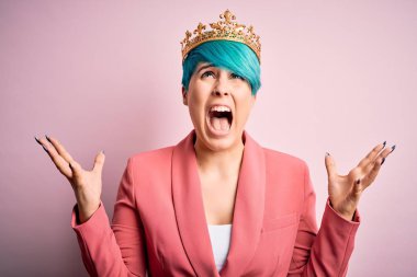 Young business woman with blue fashion hair wearing queen crown over pink isolated background crazy and mad shouting and yelling with aggressive expression and arms raised. Frustration concept.