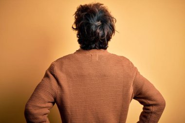 Young handsome man wearing casual shirt and sweater over isolated yellow background standing backwards looking away with arms on body