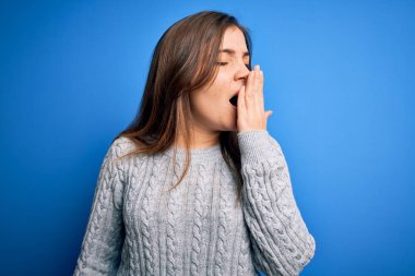 Mavi izole arka planda dikilen günlük yün süveter giyen güzel genç bir kadın ağzıyla esnemekten sıkılmış. Huzursuz ve uykulu..