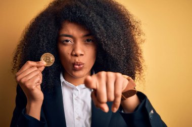 Young african american business woman with afro hair holding bitcoin over yellow background pointing with finger to the camera and to you, hand sign, positive and confident gesture from the front