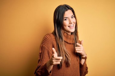 İzole edilmiş sarı arka planda dikilip mutlu ve komik yüzlü parmakları kameraya gösteren gündelik süveter giyen genç ve güzel bir kız. İyi enerji ve titreşimler.