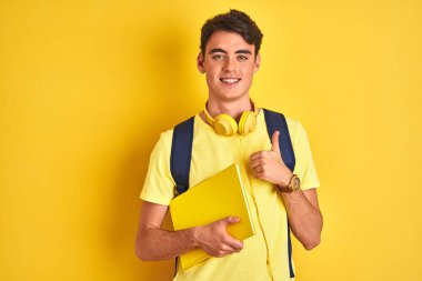 Kulaklık takan ve sırt çantası olan genç bir çocuk izole edilmiş arka planda kitap okuyor. Gülümseyerek mutlu oluyor. Tamam işareti yapıyor.