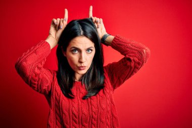 Mavi gözlü genç esmer kadın izole edilmiş kırmızı arka plan üzerine gündelik süveter giyiyor ve boynuzlarını başlarının üzerinde gezdirerek komik hareketler yapıyor.