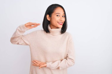 Young chinese woman wearing turtleneck sweater standing over isolated white background gesturing with hands showing big and large size sign, measure symbol. Smiling looking at the camera. Measuring concept.