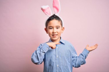Paskalya tavşanı kulakları takan küçük bir çocuk pembe arka planda hayrete düşmüş ve kameraya gülümserken eliyle ve parmağıyla işaret ediyor..