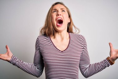 Young beautiful blonde woman wearing casual striped t-shirt over isolated white background crazy and mad shouting and yelling with aggressive expression and arms raised. Frustration concept.