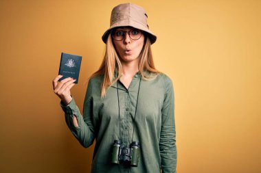 Blonde tourist woman with blue eyes on vacation holding australian passport using binoculars scared in shock with a surprise face, afraid and excited with fear expression