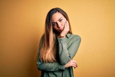 Young beautiful blonde woman with blue eyes wearing green shirt over yellow background thinking looking tired and bored with depression problems with crossed arms.