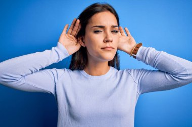Young beautiful brunette woman wearing casual sweater standing over blue background Trying to hear both hands on ear gesture, curious for gossip. Hearing problem, deaf