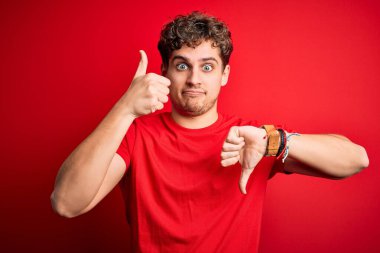 Young blond handsome man with curly hair wearing casual t-shirt over red background Doing thumbs up and down, disagreement and agreement expression. Crazy conflict