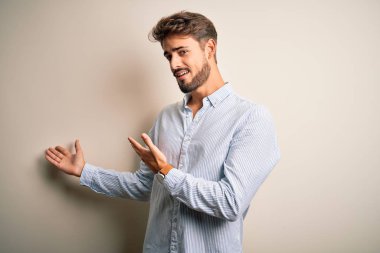 Young handsome man with beard wearing striped shirt standing over white background Inviting to enter smiling natural with open hand