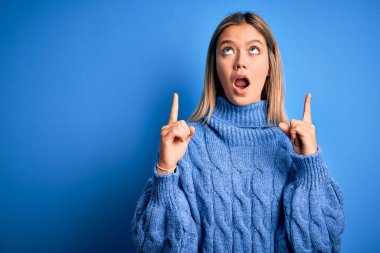 Young beautiful blonde woman wearing winter wool sweater over blue isolated background amazed and surprised looking up and pointing with fingers and raised arms.