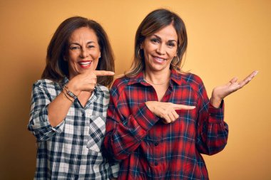 Orta yaşlı, güzel iki kız kardeş, izole edilmiş sarı arka plan üzerine gündelik tişört giyip kameraya hayret ediyor ve gülümsüyor..