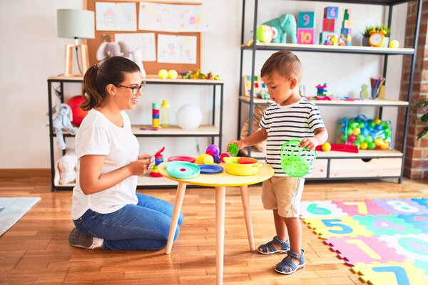 Beautiful toddler boy sitting on puzzle playing with plastic plates, fruits and vegetables at kindergarten