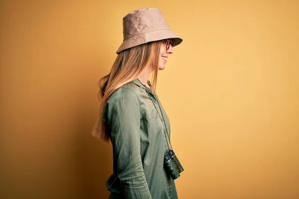 Beautiful blonde explorer woman with blue eyes wearing hat and glasses using binoculars looking to side, relax profile pose with natural face with confident smile.